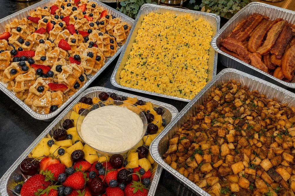 Chef-prepared trays of waffles, eggs, bacon, potatoes, and a fresh fruit platter, ready to serve